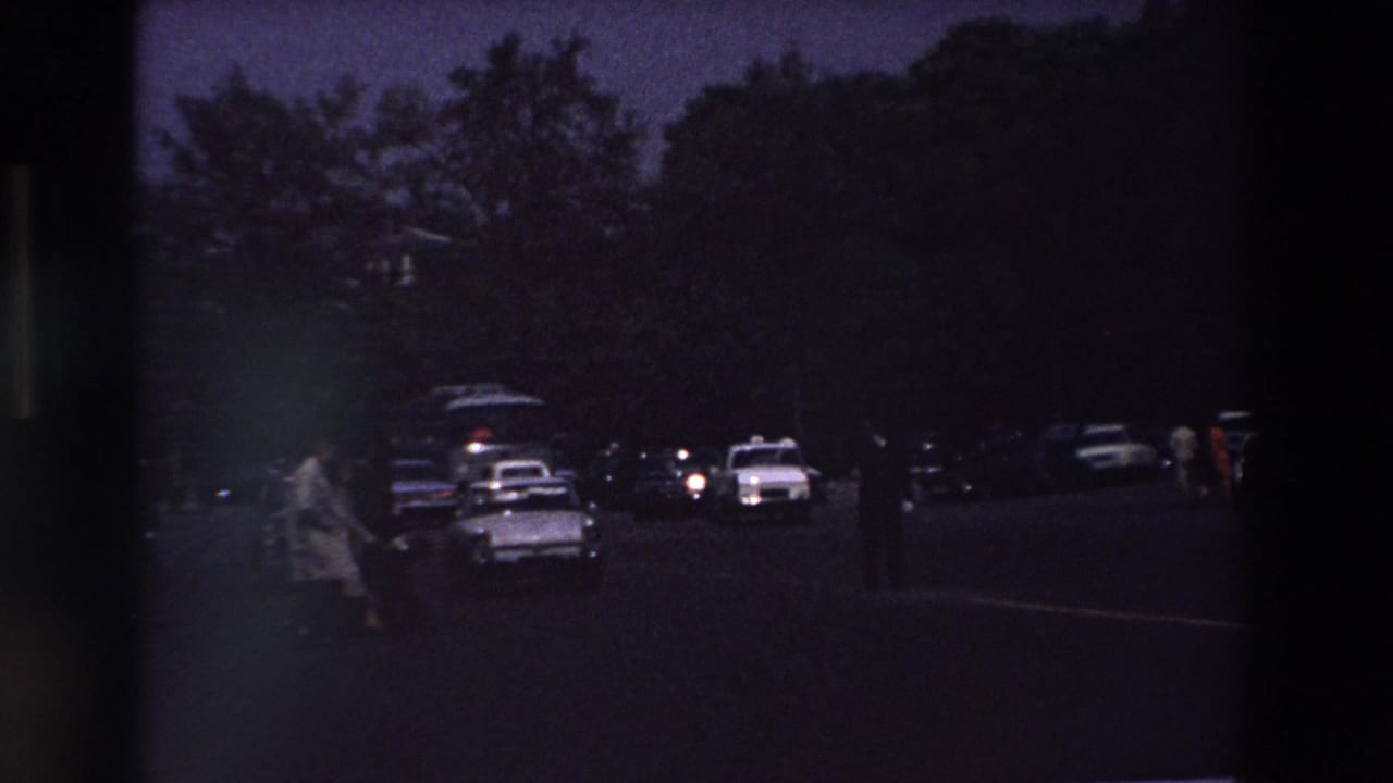 Dark 1969 Paris parking-lot scene with silhouetted trees, several classic cars, and a person walking near a white taxi-like vehicle.
