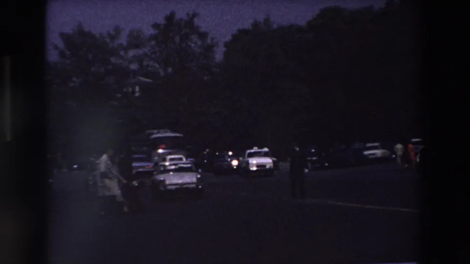 Dark 1969 Paris parking-lot scene with silhouetted trees, several classic cars, and a person walking near a white taxi-like vehicle.