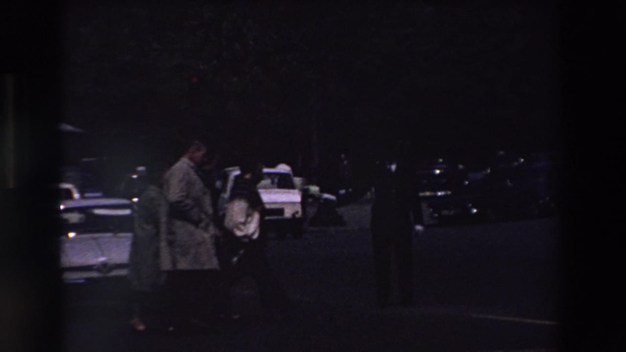 Two people in period coats appear near parked cars in a low-light Paris parking lot, with a white taxi-like car and roof light visible behind them.