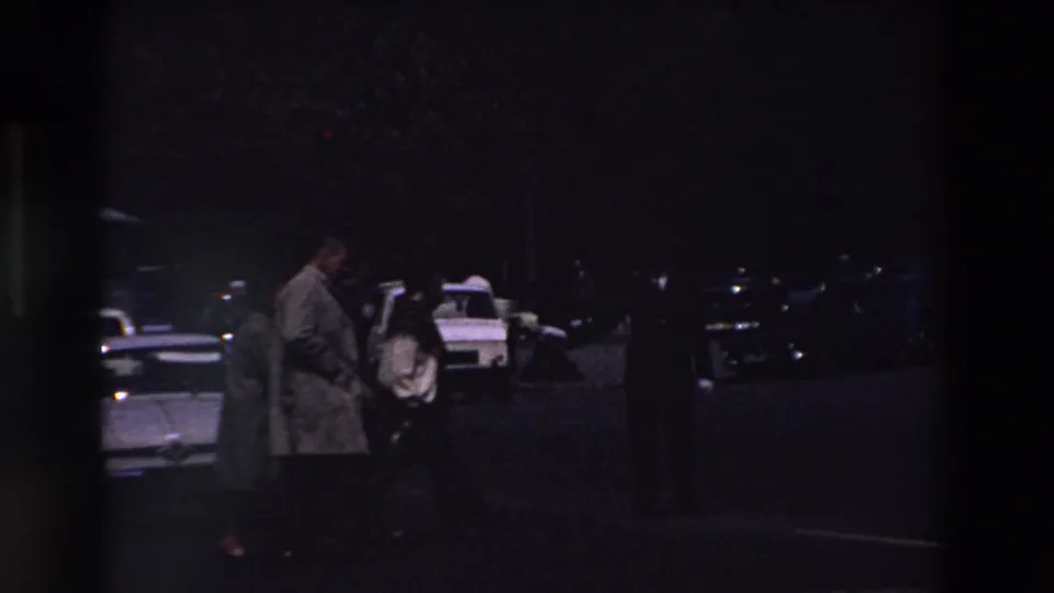 Two people in period coats appear near parked cars in a low-light Paris parking lot, with a white taxi-like car and roof light visible behind them.
