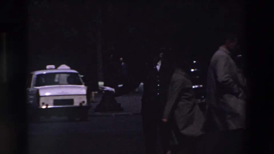 Low-light 8mm frame from Paris showing pedestrians in coats crossing or standing near a white taxi-like car among dark parked automobiles.