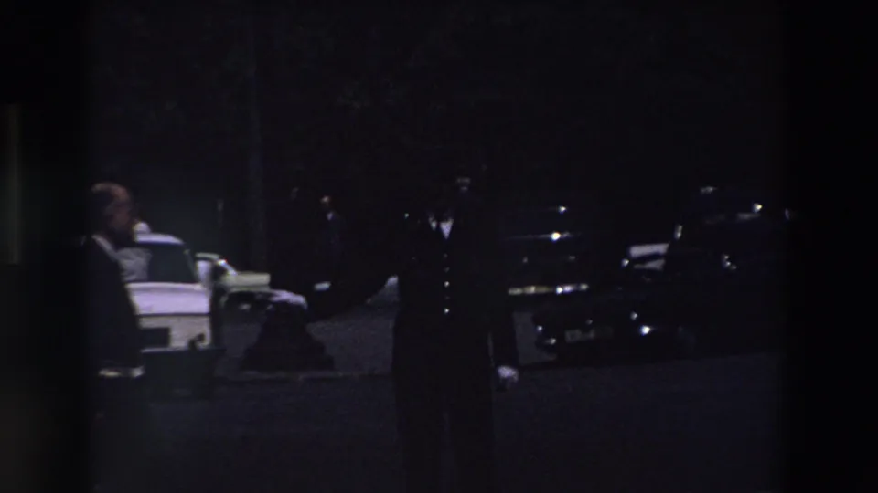 A suited figure stands in a dark Paris parking-lot scene with parked cars, a white vehicle, and deep shadows from the nighttime 8mm exposure.