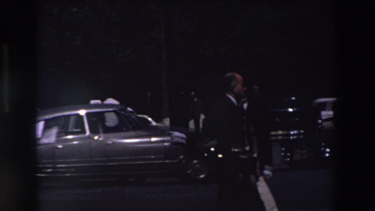 Nighttime Paris street scene from 1969 home-movie footage, showing a suited man walking near parked mid-century cars under very low light.