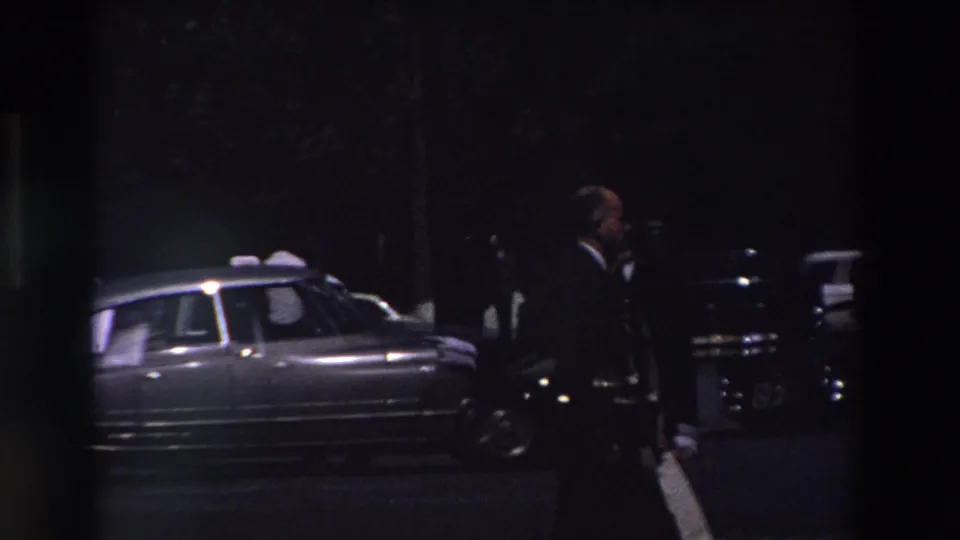 Nighttime Paris street scene from 1969 home-movie footage, showing a suited man walking near parked mid-century cars under very low light.