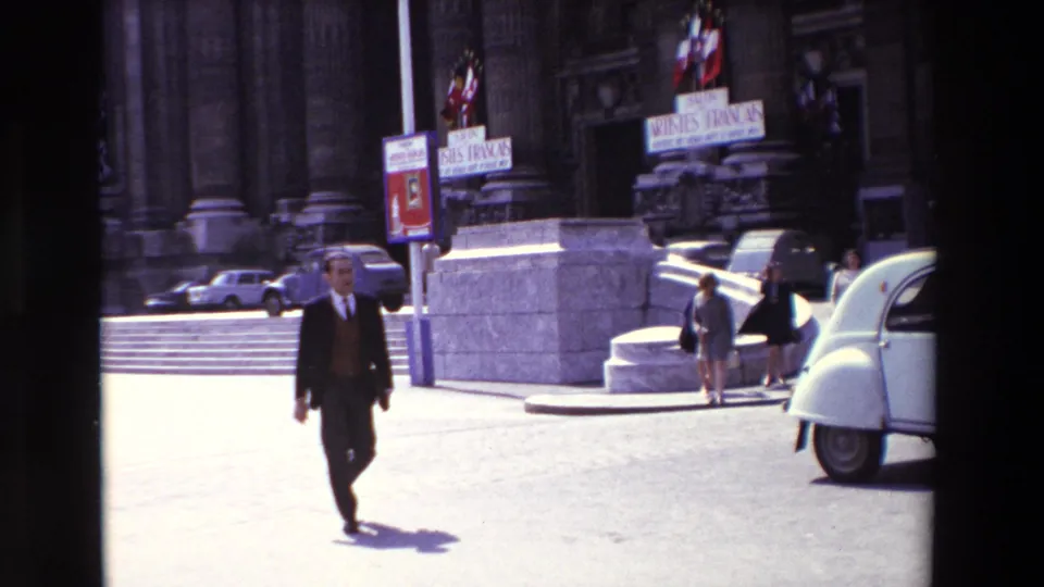 Daylight Paris plaza scene from 1969 footage, with a suited man walking toward the camera near a white Citroen 2CV-style car, stone steps, French flags, and Artistes Français signage.