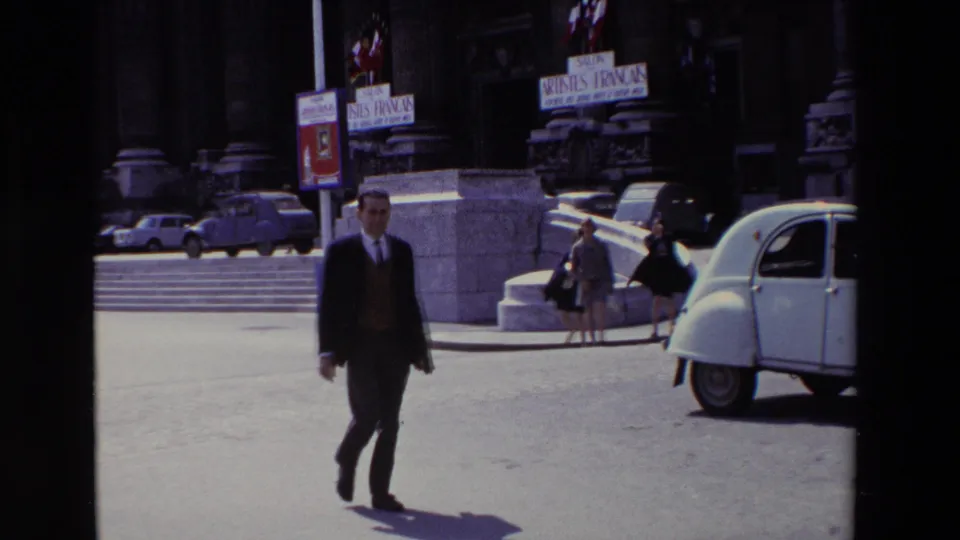 1969 Paris daylight frame showing a suited man walking across open pavement with a white Citroen 2CV-style car, pedestrians, steps, flags, and Artistes Français signage behind him.