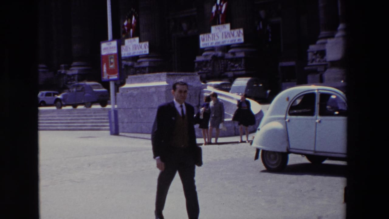 Paris, France, 1969 home-movie frame showing a suited man in the foreground with a white Citroën 2CV, pedestrians, stone architecture, and Salon des Artistes Français signs behind him.