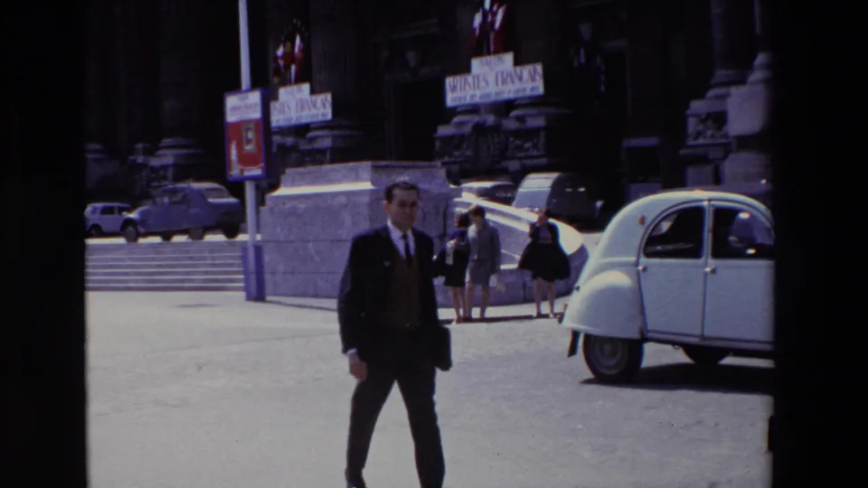 Paris, France, 1969 home-movie frame showing a suited man in the foreground with a white Citroën 2CV, pedestrians, stone architecture, and Salon des Artistes Français signs behind him.