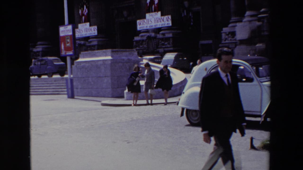 Suited man walks out of frame beside a parked white car as pedestrians stand near stone steps.