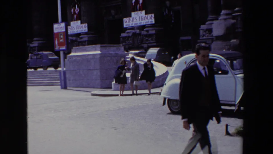 Paris, France, 1969 frame with a man in a dark suit at the right edge, a white Citroën 2CV, pedestrians near a stone entrance, and visible Salon des Artistes Français signage.
