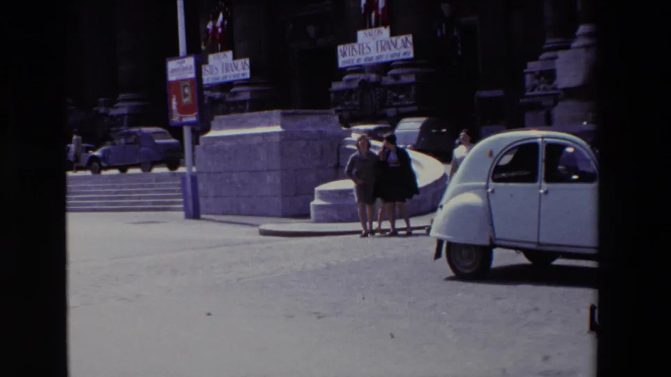 Paris, France, 1969 home-movie frame showing women in 1960s attire near broad stone steps, exhibition posters, parked cars, and a white Citroën 2CV in the foreground.