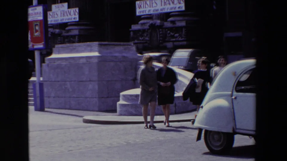 Paris, France, 1969 frame showing two women walking arm in arm near a white Citroën 2CV, with other pedestrians, stone steps, and Salon des Artistes Français signage in the background.