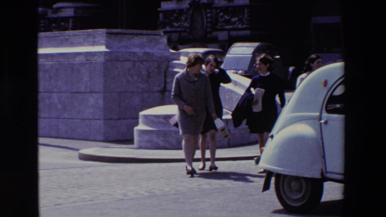 In supported 1969 Paris footage, several adult women walk together near stone steps and a white Citroen 2CV on a bright city street.