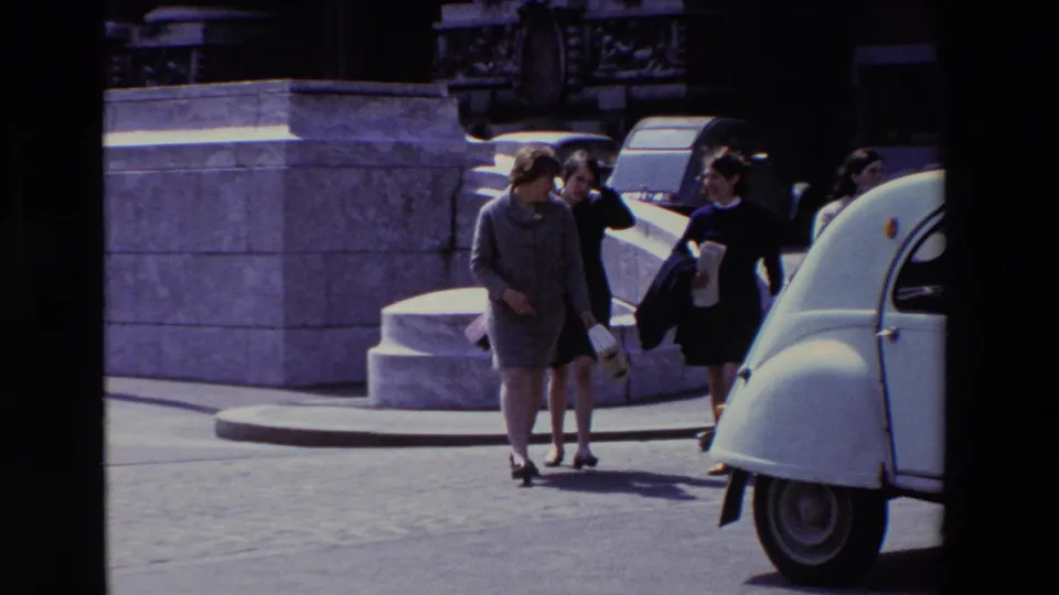 In supported 1969 Paris footage, several adult women walk together near stone steps and a white Citroen 2CV on a bright city street.