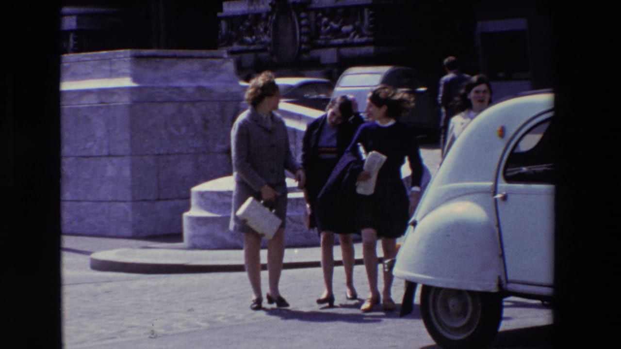 Adult women in coats and skirts pause or walk together on a Paris sidewalk, with a white Citroen 2CV cropped at the right edge.