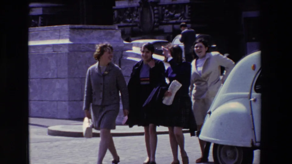Three women walk toward the foreground on a Paris sidewalk while another woman follows behind, with stone architecture and a white Citroen 2CV nearby.