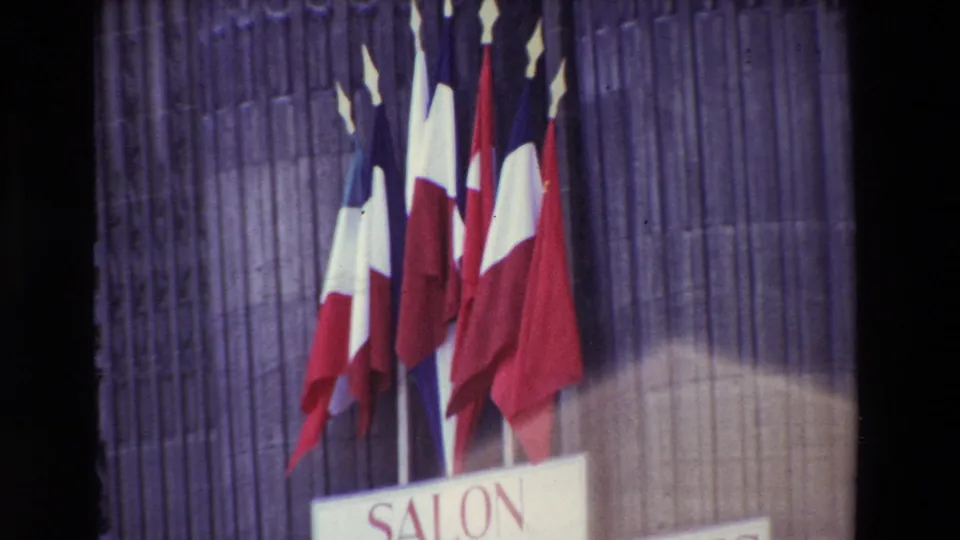 Paris, France in 1969: a tight 8mm frame shows multiple French flags and part of the Salon des Artistes Français signage against the ribbed stone facade, with a blurred passerby or foreground head at the lower right.
