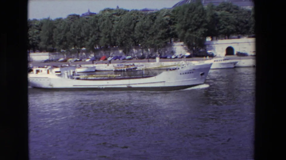 1969 Paris home-movie frame showing a white yacht on the likely Seine with river water, trees, parked cars, and a stone embankment behind it.