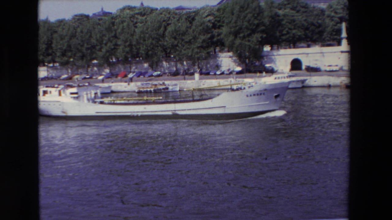 In Paris, France according to the clip metadata, a white cargo-style boat marked SAMBRE moves along the river in front of a stone embankment, parked cars, trees, and an arched wall opening.