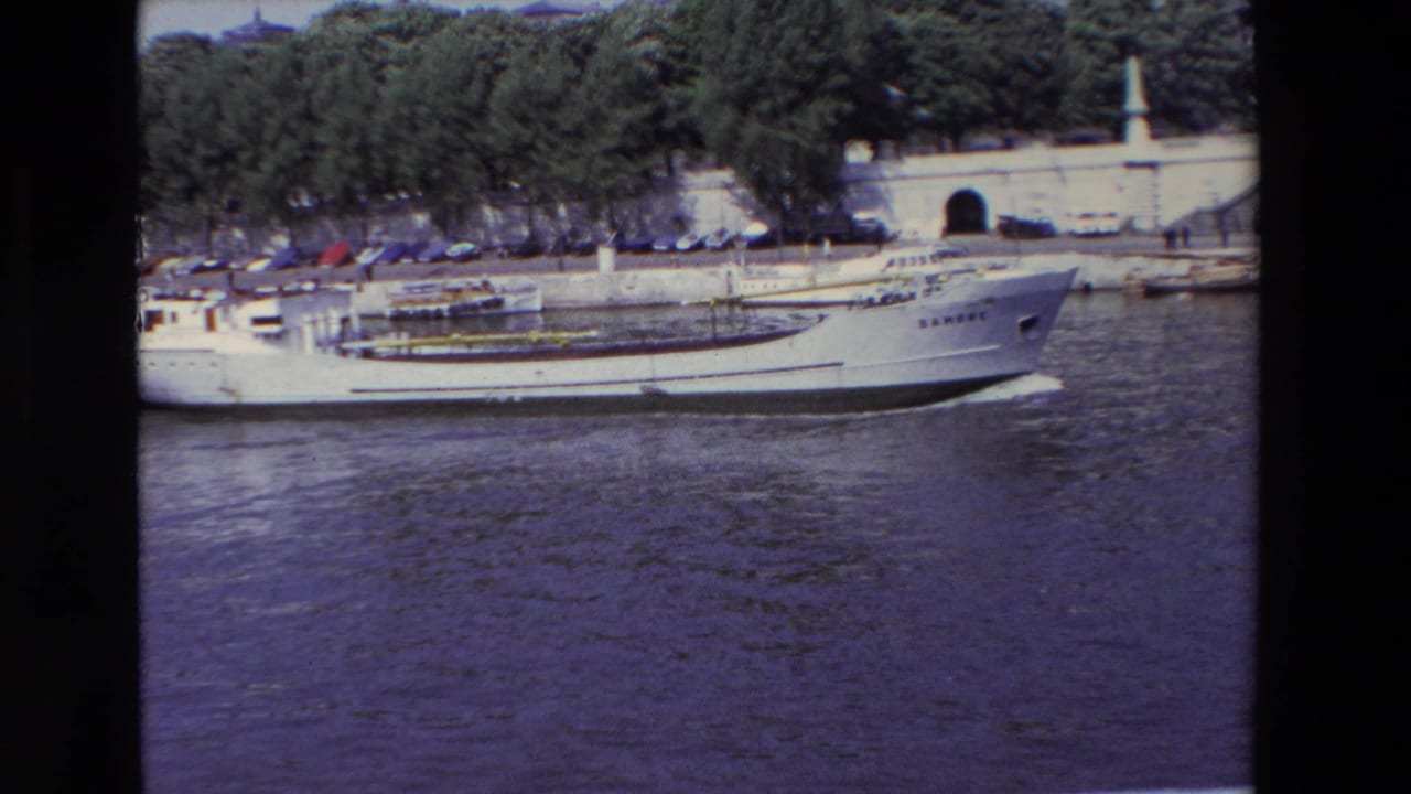 The white boat continues across the Paris river scene, with its bow near the right side of frame and a tree-lined stone embankment, parked cars, and arched opening behind it.