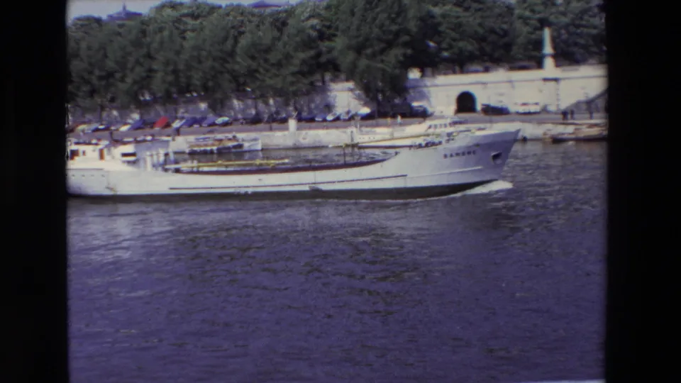 The white boat continues across the Paris river scene, with its bow near the right side of frame and a tree-lined stone embankment, parked cars, and arched opening behind it.