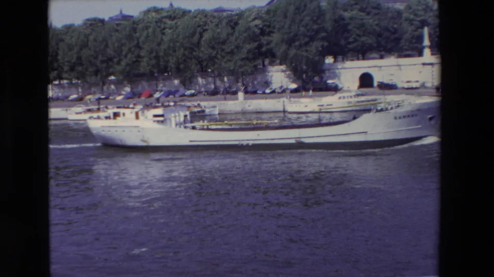 A side view shows the white SAMBRE boat moving steadily along the river in Paris, with wake on the water and a tree-lined embankment with parked cars in the background.