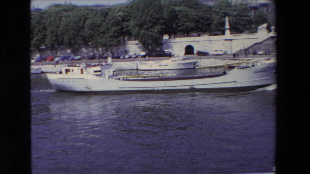 1969 Paris 8mm frame showing a white river transport boat on the Seine with a stone riverside structure, parked cars, and trees behind it.