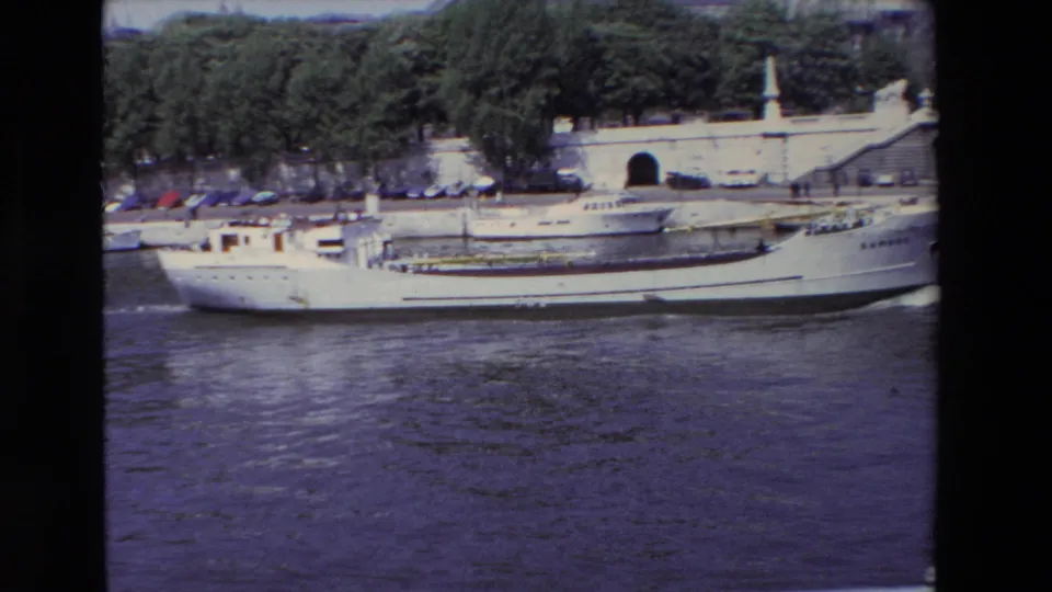 1969 Paris 8mm frame showing a white river transport boat on the Seine with a stone riverside structure, parked cars, and trees behind it.