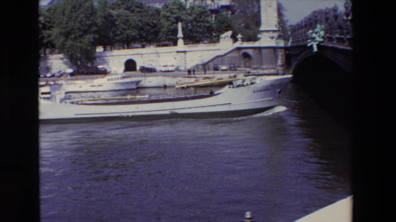 1969 Paris 8mm frame of a white boat traveling on the Seine below a stone bridge or quay structure with parked cars and leafy trees above.