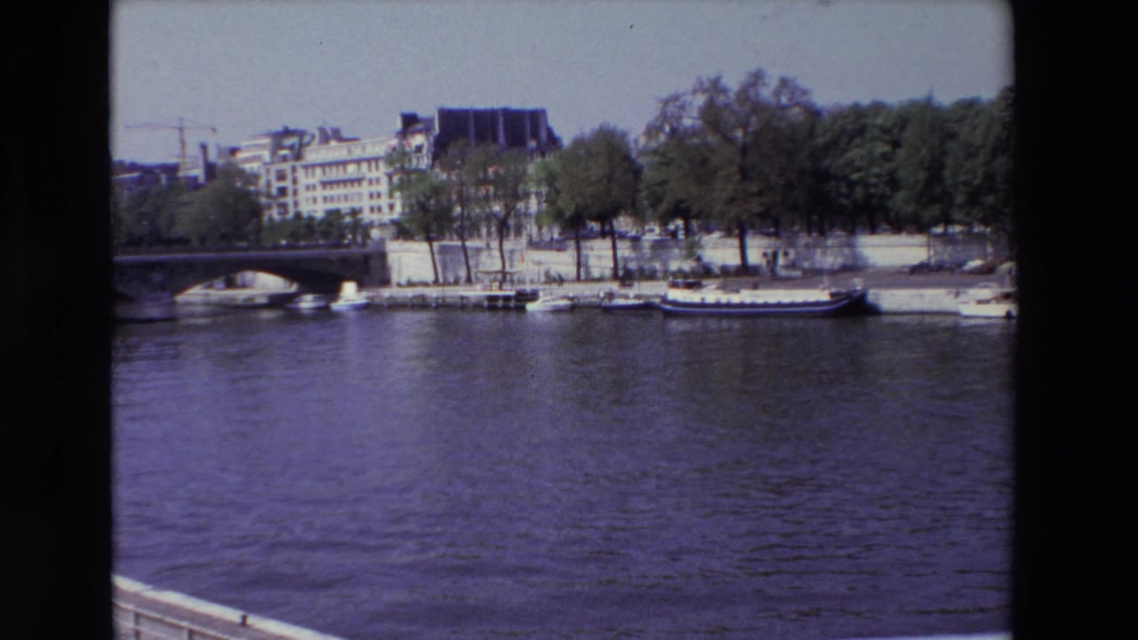 1969 Paris 8mm frame looking across the Seine toward moored boats, a stone bridge, tree-lined embankment, and older riverside buildings.