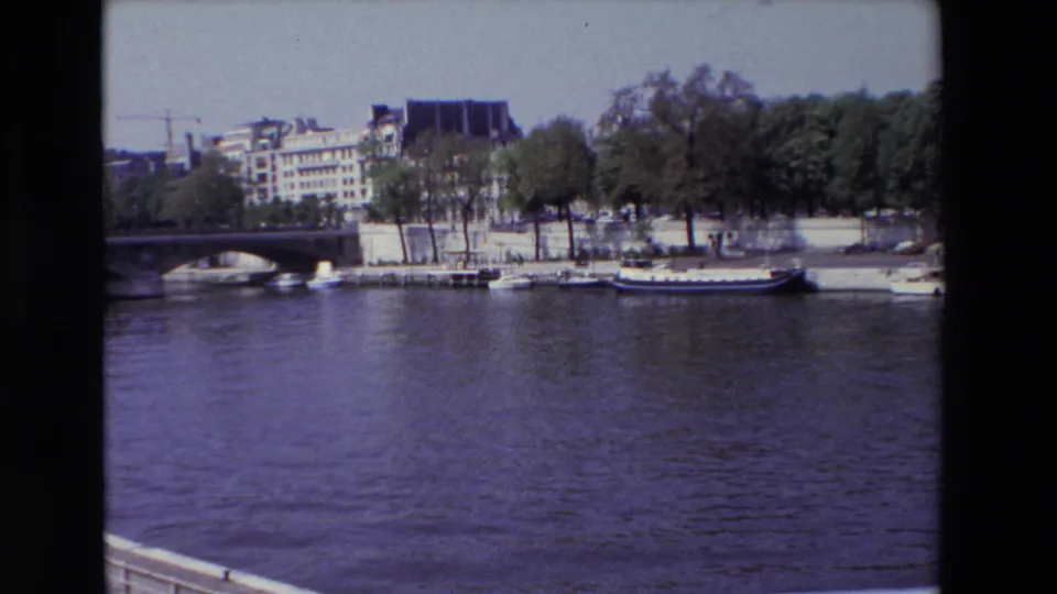 1969 Paris 8mm frame looking across the Seine toward moored boats, a stone bridge, tree-lined embankment, and older riverside buildings.