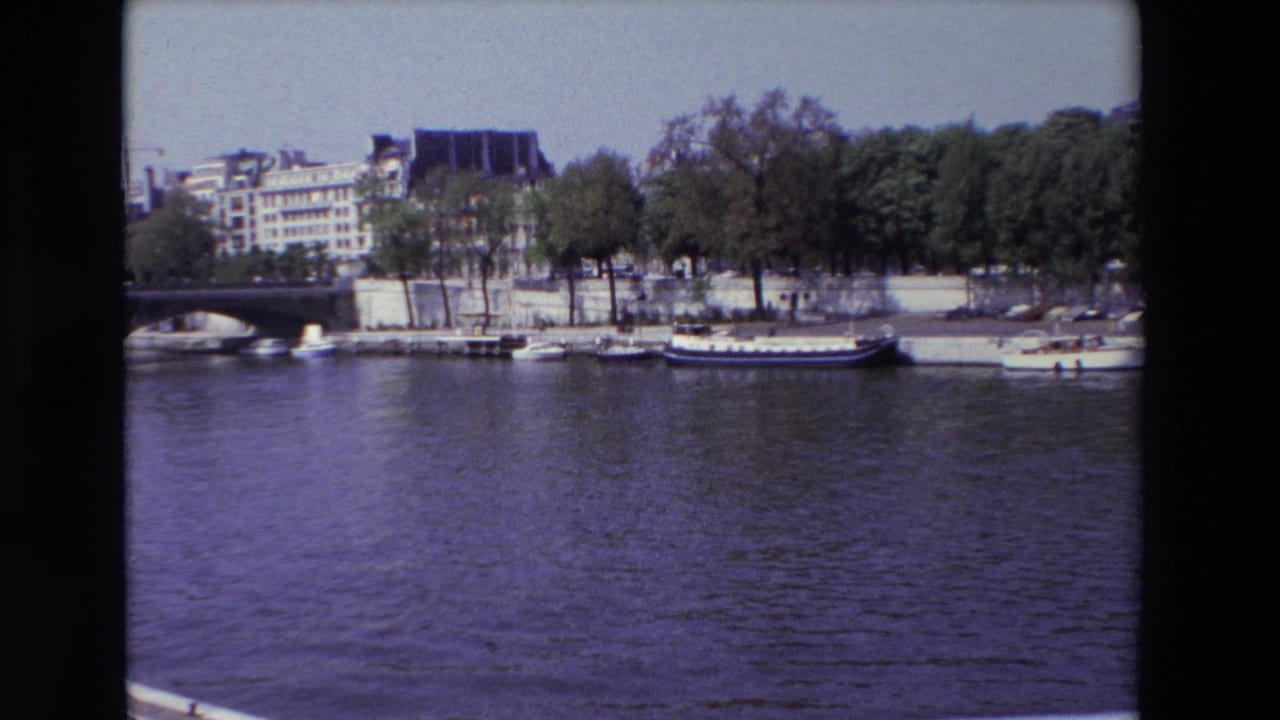 1969 Paris home-movie frame showing the Seine in the foreground with boats tied along the quay, trees, a partial bridge, and city buildings beyond.