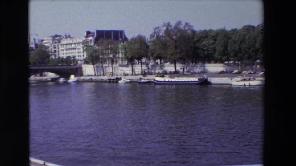 1969 Paris home-movie frame showing the Seine in the foreground with boats tied along the quay, trees, a partial bridge, and city buildings beyond.
