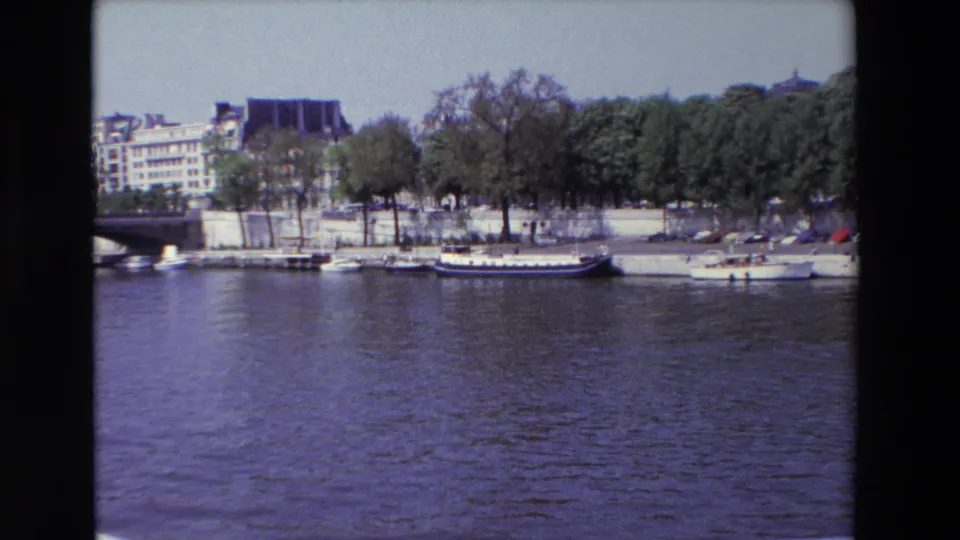 1969 Paris 8mm frame showing boats along the Seine embankment, parked cars under trees, and older buildings in the background.