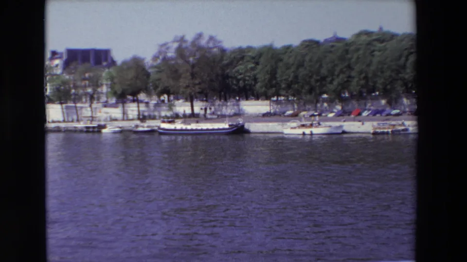 1969 Paris home-movie frame with the Seine in the foreground, boats lined along the stone quay, trees and parked cars on the bank, and buildings behind them.