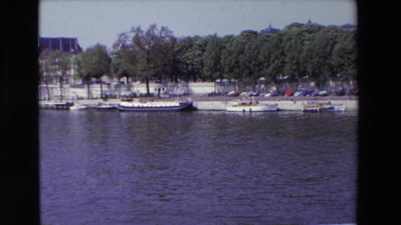 1969 Paris home-movie frame showing moored boats beside a tree-lined quay, with rippling river water across the foreground.