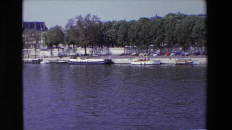 1969 Paris home-movie frame showing moored boats beside a tree-lined quay, with rippling river water across the foreground.