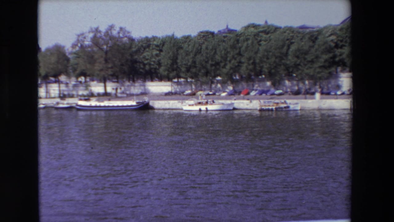 1969 Paris riverside view with small craft, a larger moored vessel, parked cars, and dense trees behind the quay.