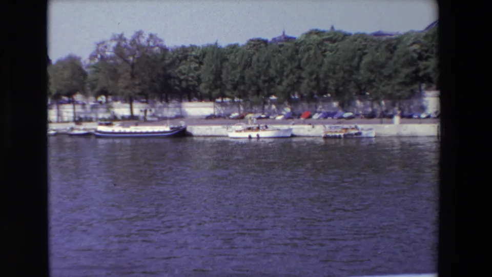 1969 Paris riverside view with small craft, a larger moored vessel, parked cars, and dense trees behind the quay.