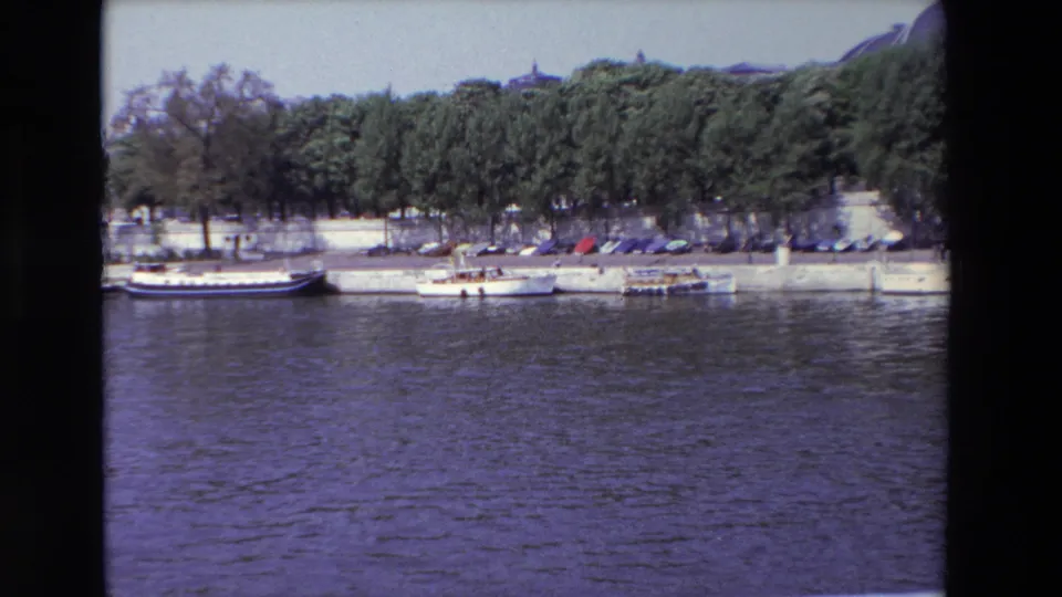 1969 Paris home-movie frame looking across the river toward a stone embankment, moored boats, parked cars, and leafy trees.