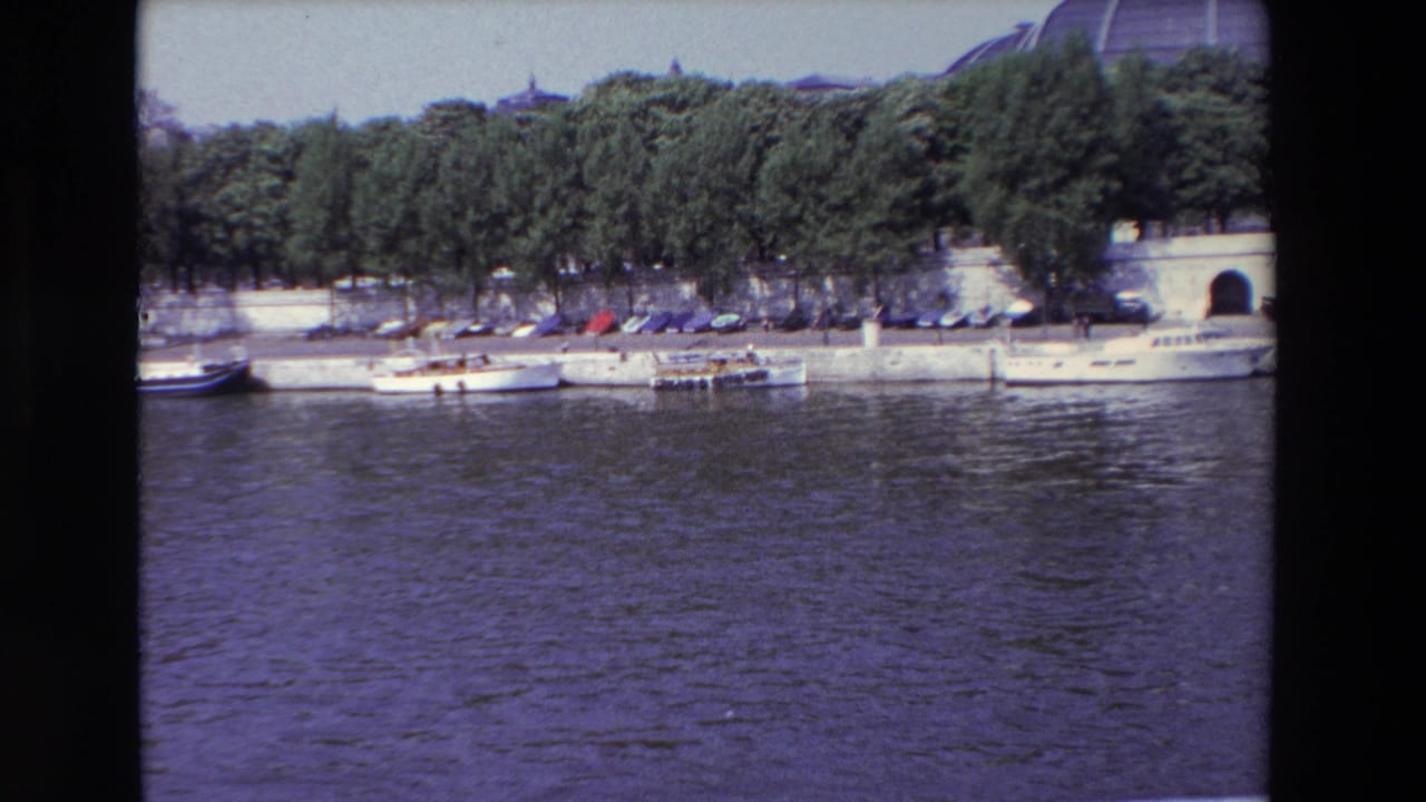 1969 Paris home-movie frame showing rippling river water, small white boats tied along a concrete quay, trees, parked cars, and the edge of a domed building in the background.