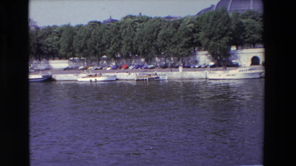 1969 Paris home-movie frame showing rippling river water, small white boats tied along a concrete quay, trees, parked cars, and the edge of a domed building in the background.