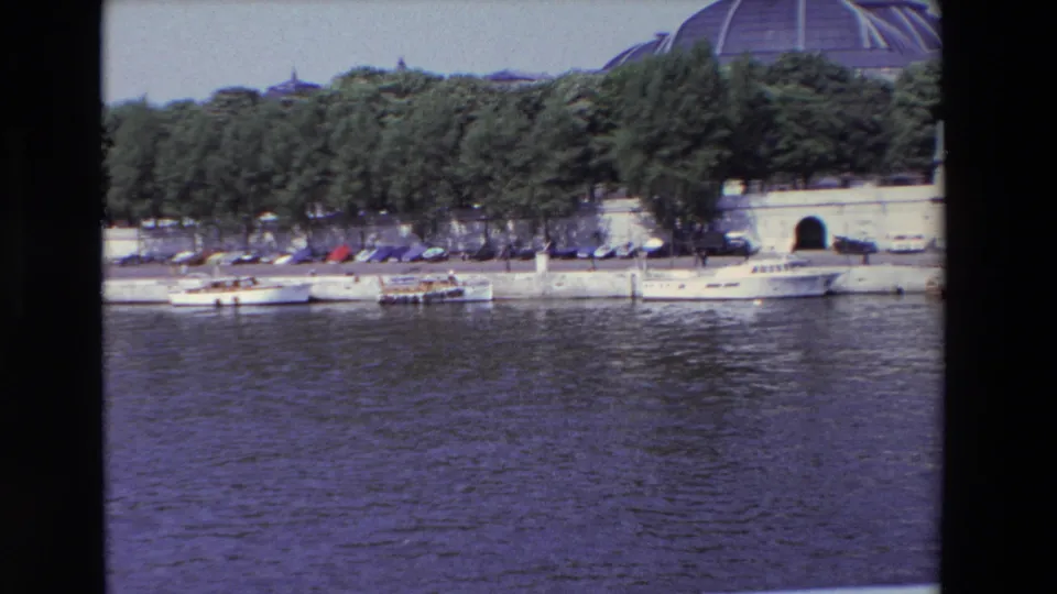 1969 Paris riverside frame with moored boats beside a concrete embankment, dense trees, a visible tunnel entrance, parked automobiles, and a domed structure partly above the treeline.