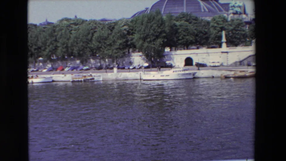 1969 Paris frame showing the camera pan farther along the riverbank, with white boats at the quay, parked cars behind trees, a dark tunnel opening, and a domed roofline in the upper background.
