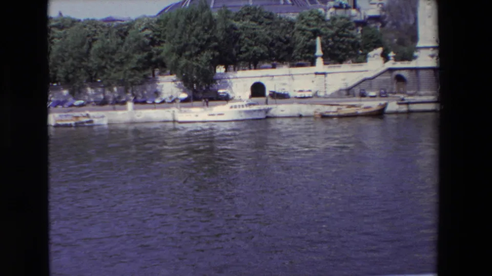 1969 Paris home-movie frame with broad water foreground, a line of boats tied to the quay, trees and parked cars along the embankment, a tunnel entrance, steps, and part of a domed building above.