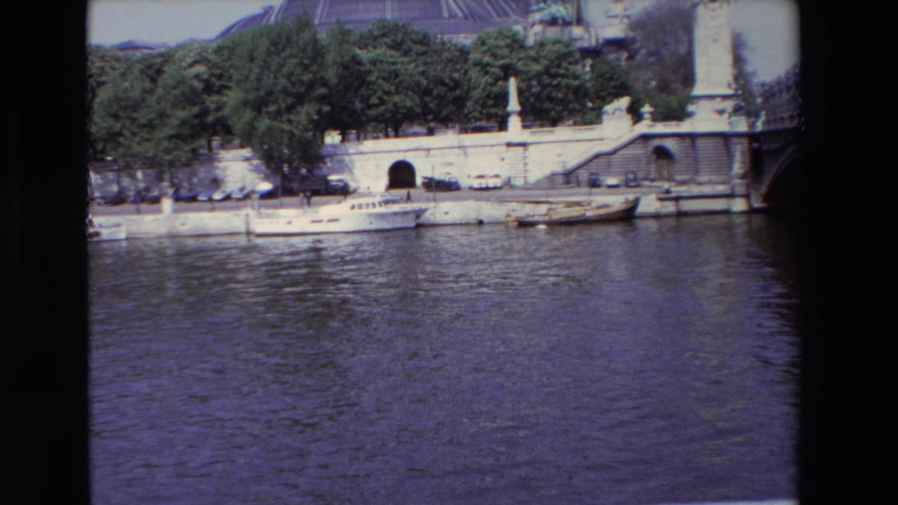 1969 Paris home-movie frame showing the Seine, a stone quay with small boats and parked cars, trees, and the Grand Palais dome in the background.