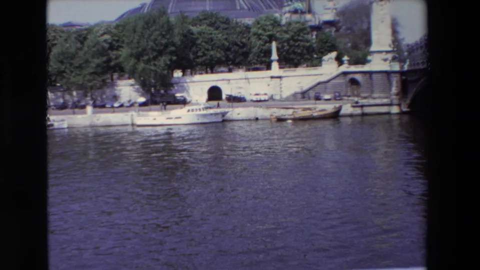 1969 Paris home-movie frame showing the Seine, a stone quay with small boats and parked cars, trees, and the Grand Palais dome in the background.