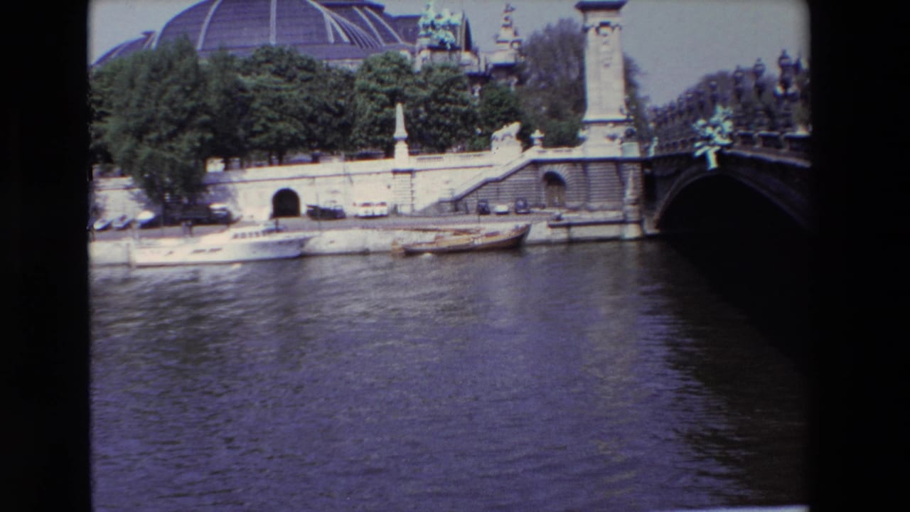 1969 Paris frame looking across the Seine toward moored boats, parked cars along the quay, trees, the Grand Palais dome, and the dark edge of a low bridge.