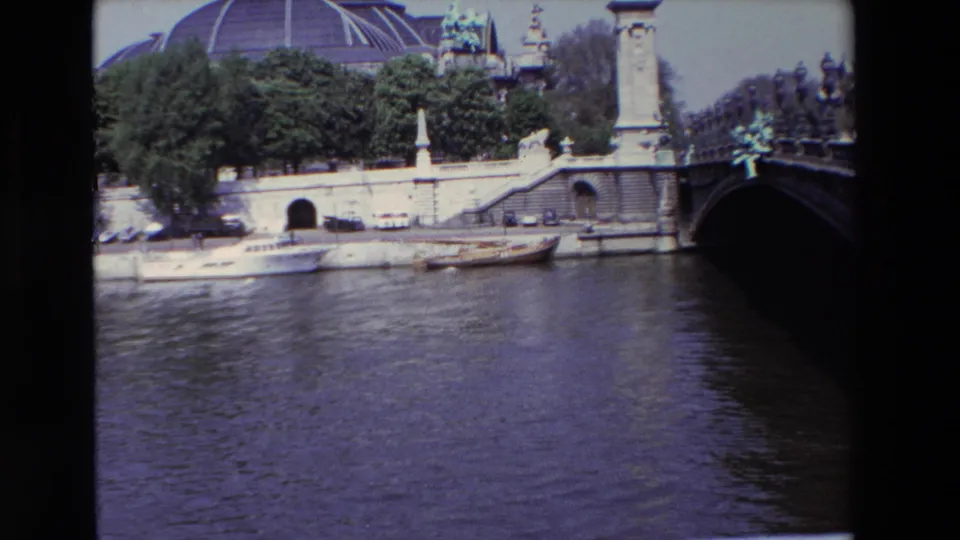 1969 Paris frame looking across the Seine toward moored boats, parked cars along the quay, trees, the Grand Palais dome, and the dark edge of a low bridge.