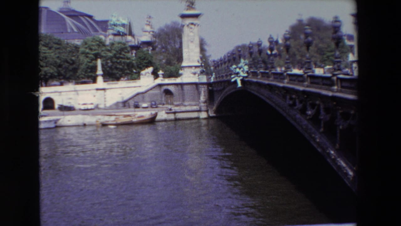Low bridge crosses the Seine with ornate railings, stone supports, boats, trees, and Grand Palais roof behind.